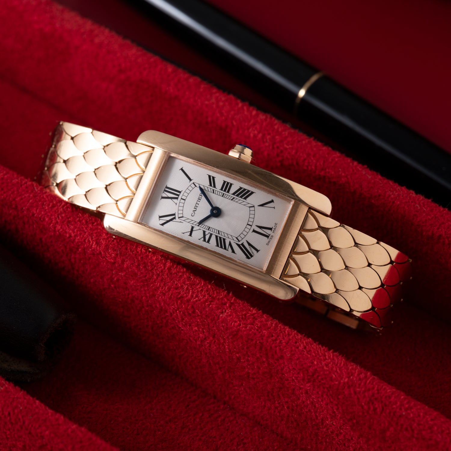 A rectangular ladies' Cartier Tank Americaine in 18K rose gold with a silver guilloché dial and Roman numerals lies on red fabric, with portions of a black pen and leather case visible nearby.