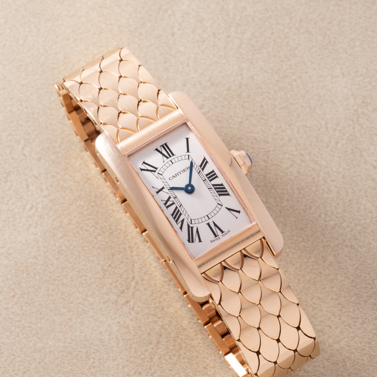 A Cartier Tank Americaine ladies wristwatch in 18K rose gold with a silver guilloché dial, Roman numerals, blue hands, and a gold scale-pattern bracelet by Cartier, shown on a beige textured surface.
