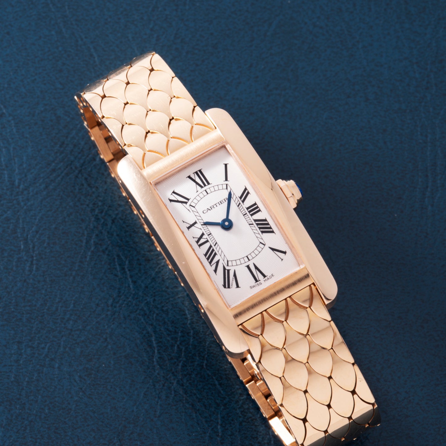 A Cartier Tank Americaine ladies wristwatch in 18K rose gold, featuring a rectangular face with Roman numerals and a patterned gold band, is displayed on a dark textured surface.