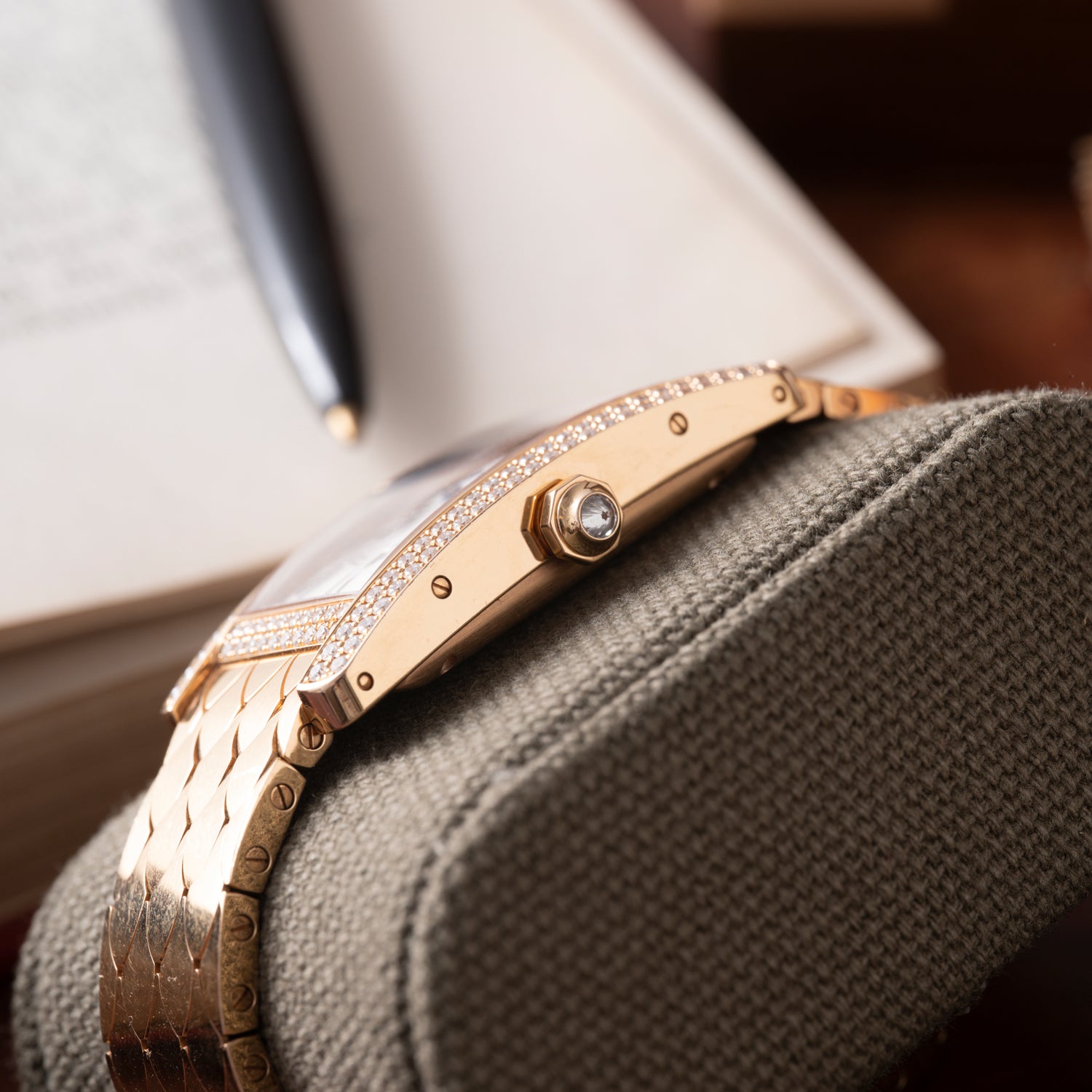 A close-up of the Cartier Tank Americaine Automatic Diamonds watch in 18K rose gold, featuring a diamond-set bezel and crown, rests on a textured cushion with an open book and black pen blurred in the background.