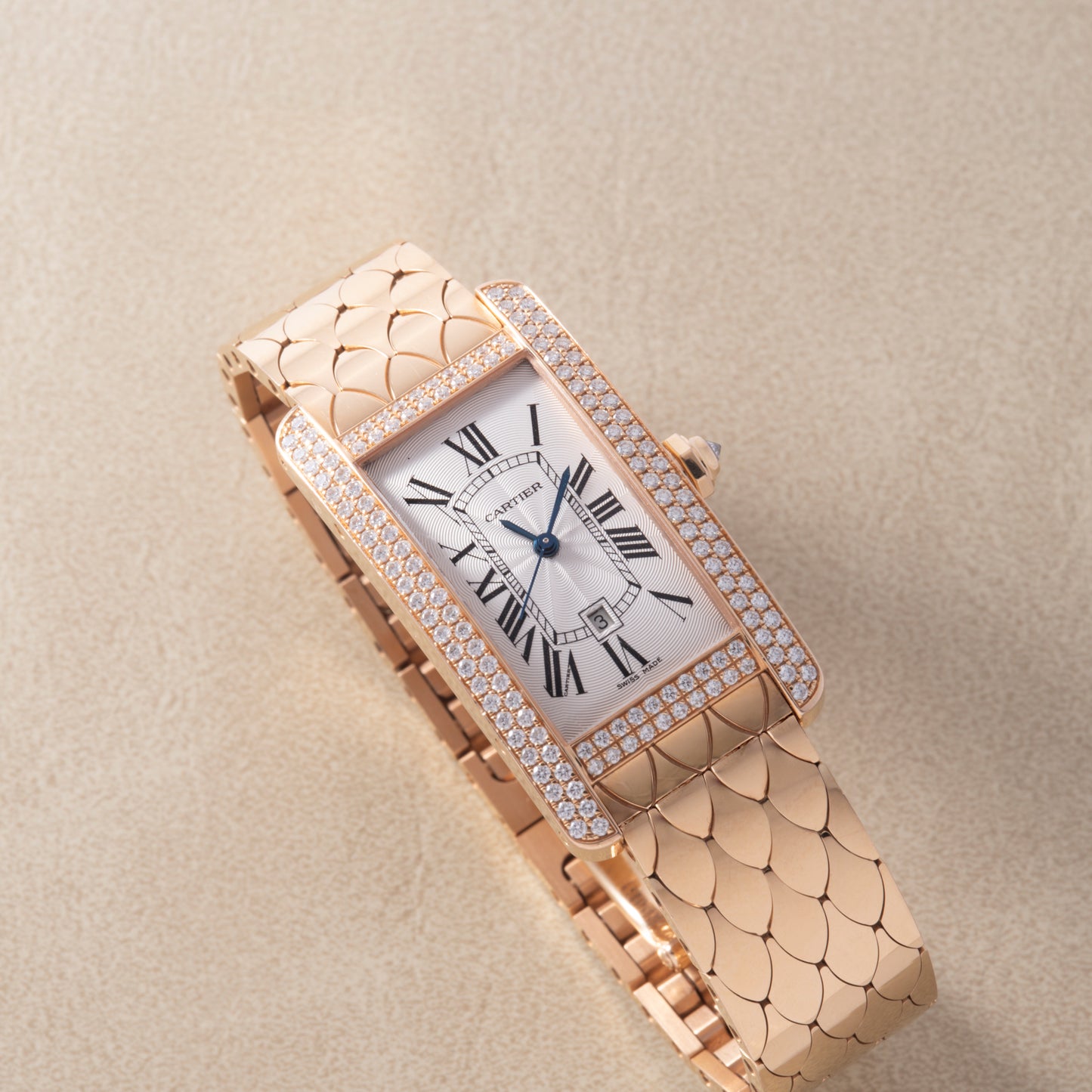 The Cartier Tank Americaine Automatic Diamonds watch by Cartier features a rectangular 18K rose gold case, diamond accents, Roman numerals on a white dial, and a gold bracelet, displayed on a beige textured surface.