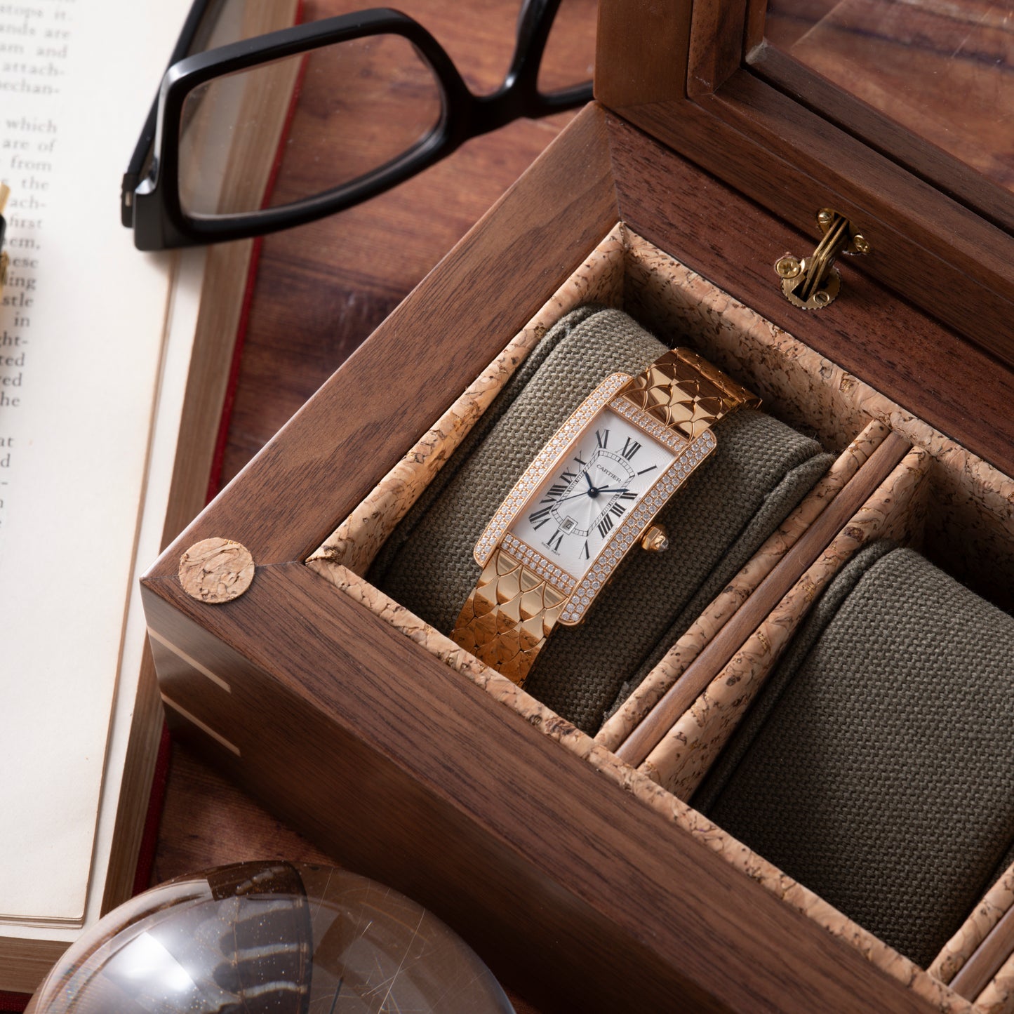 A Cartier Tank Americaine Automatic Diamonds watch by Cartier with a rectangular face is displayed in a wooden box. Next to it are black glasses, an open book, and a glass sphere on the table.
