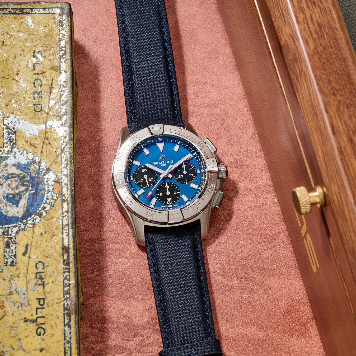 A Breitling Avenger B01 Chronograph 42 from Breitling, featuring a silver case, blue dial, and navy strap, lies on brown leather beside a vintage yellow cigar tin and a wooden box.