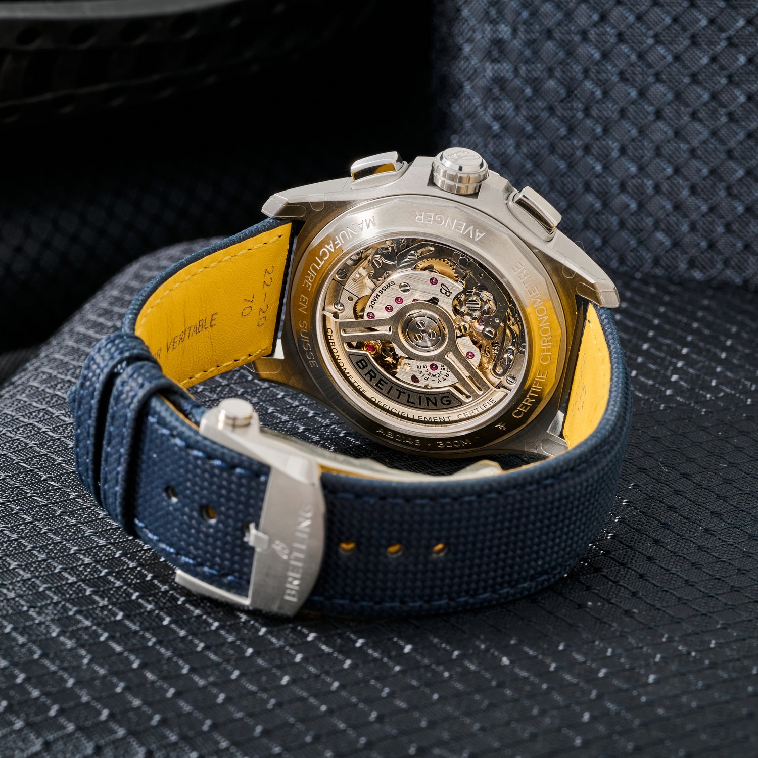 A close-up of the Breitling Avenger B01 Chronograph 42 reveals its exposed automatic movement and intricate gears, paired with a blue textured strap with yellow lining, resting on a grey textured surface.