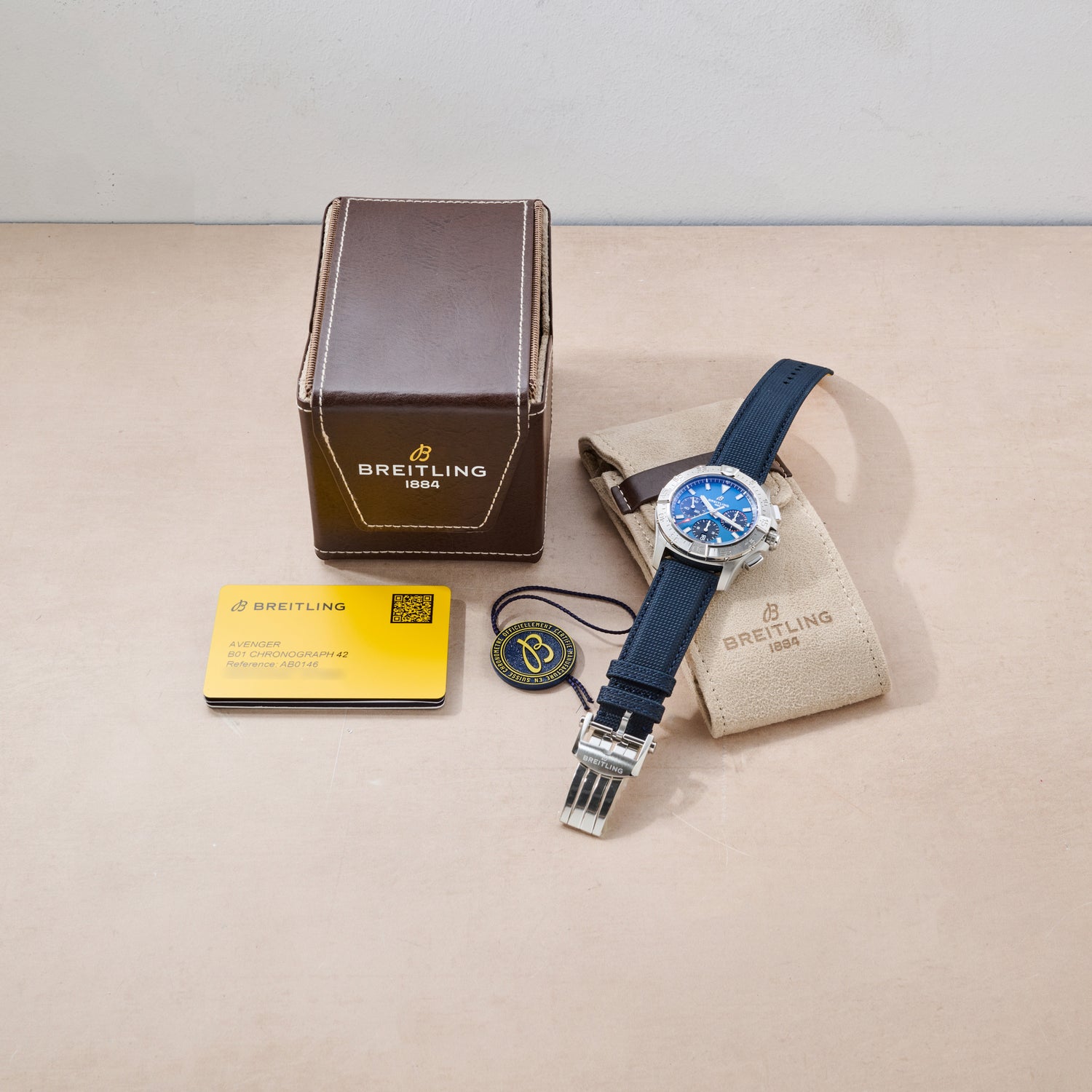 A Breitling Avenger B01 Chronograph 42 with a blue dial and black strap is shown beside its brown box, warranty card, beige pouch, and branded tag on a light beige surface.