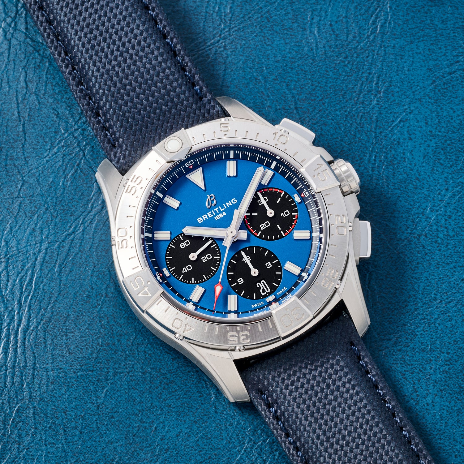 A Breitling Avenger B01 Chronograph 42 in silver with a blue dial, three black subdials, and a blue textured strap is displayed on a blue leather surface.