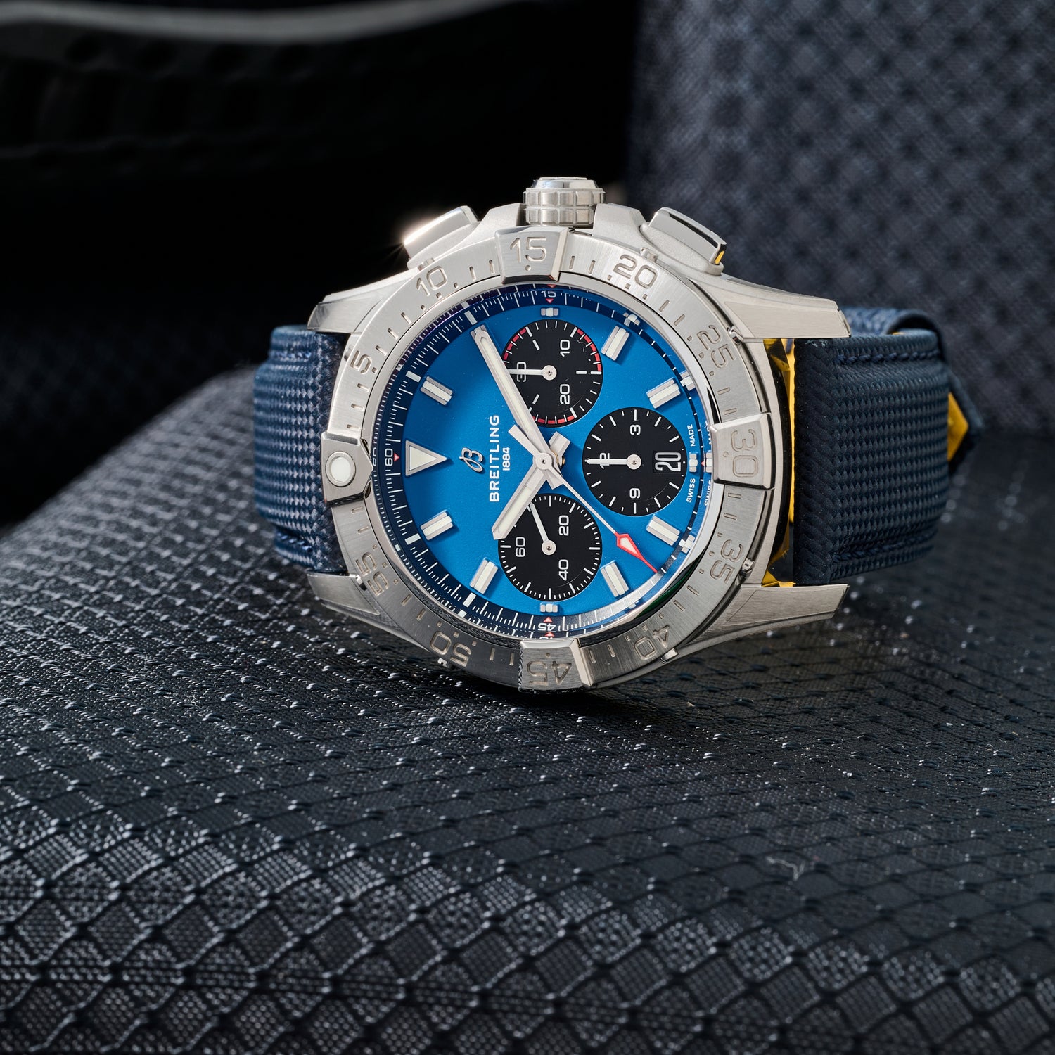 A close-up of the Breitling Avenger B01 Chronograph 42 aviation watch showcases its blue dial, silver bezel, black chronograph subdials, and textured blue strap as it rests on a patterned dark surface.