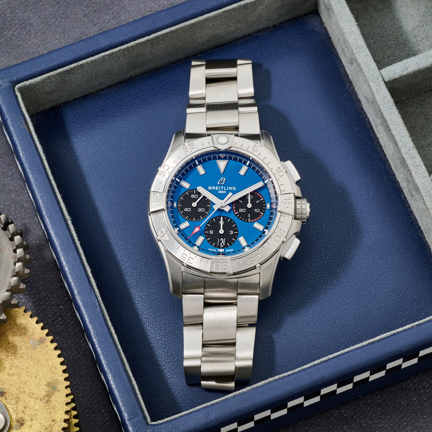 A Breitling Avenger B01 Chronograph 42 stainless steel watch with blue and black dial sits in a blue box beside a brass gear and an empty compartment.