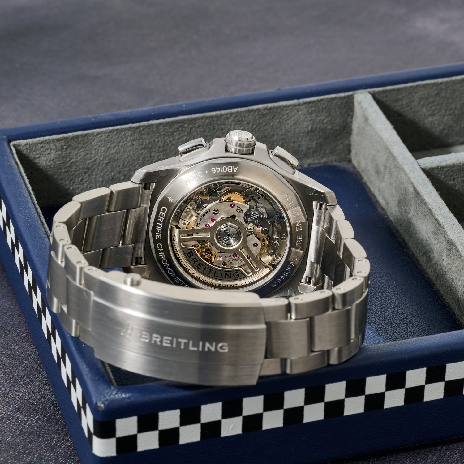A close-up of a Breitling Avenger B01 Chronograph 42 by Breitling displays its COSC-certified automatic movement with the case back facing up, resting on a blue box with checkered edges and empty gray compartments.