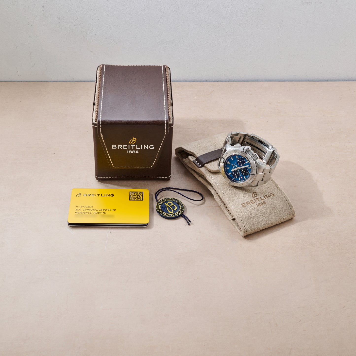 A Breitling Avenger B01 Chronograph 42 with a blue dial is shown beside a tan fabric case, brown Breitling box, yellow certificate card, and round Breitling tag on a light beige surface.