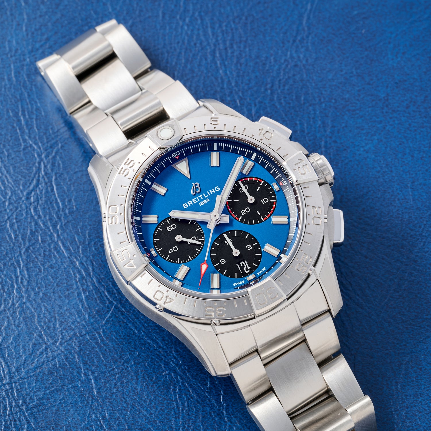 A Breitling Avenger B01 Chronograph 42 with a blue dial, three black subdials, and a stainless steel bracelet rests on a textured blue surface. This Breitling model features a COSC-certified automatic movement.