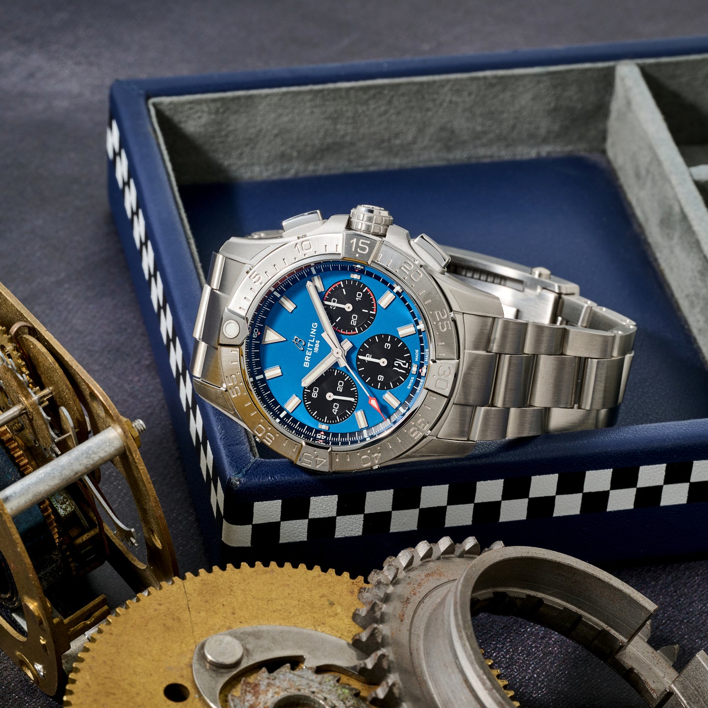 A Breitling Avenger B01 Chronograph 42 with a blue and black dial sits on a blue tray edged in black and white checkers, surrounded by metal gears in the foreground.