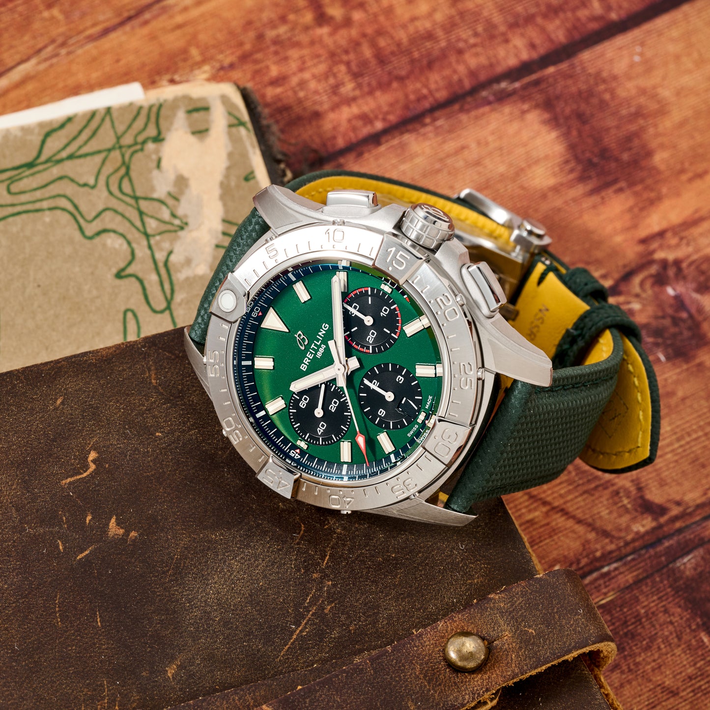 A silver Breitling Avenger B01 Chronograph 42 with a green dial and dark green strap rests on brown leather, alongside a vintage map and a wooden background.