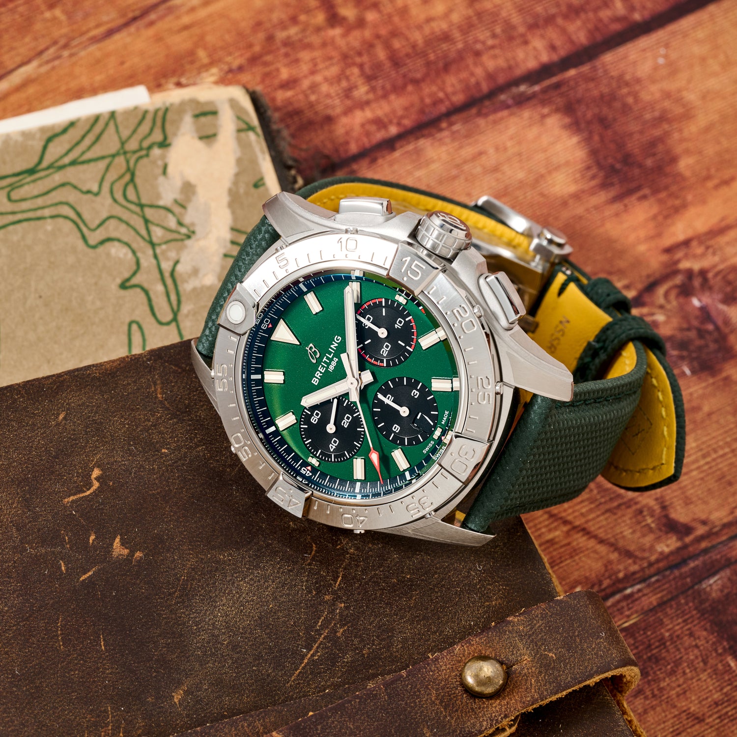 A silver Breitling Avenger B01 Chronograph 42 with a green dial and dark green strap rests on brown leather, alongside a vintage map and a wooden background.
