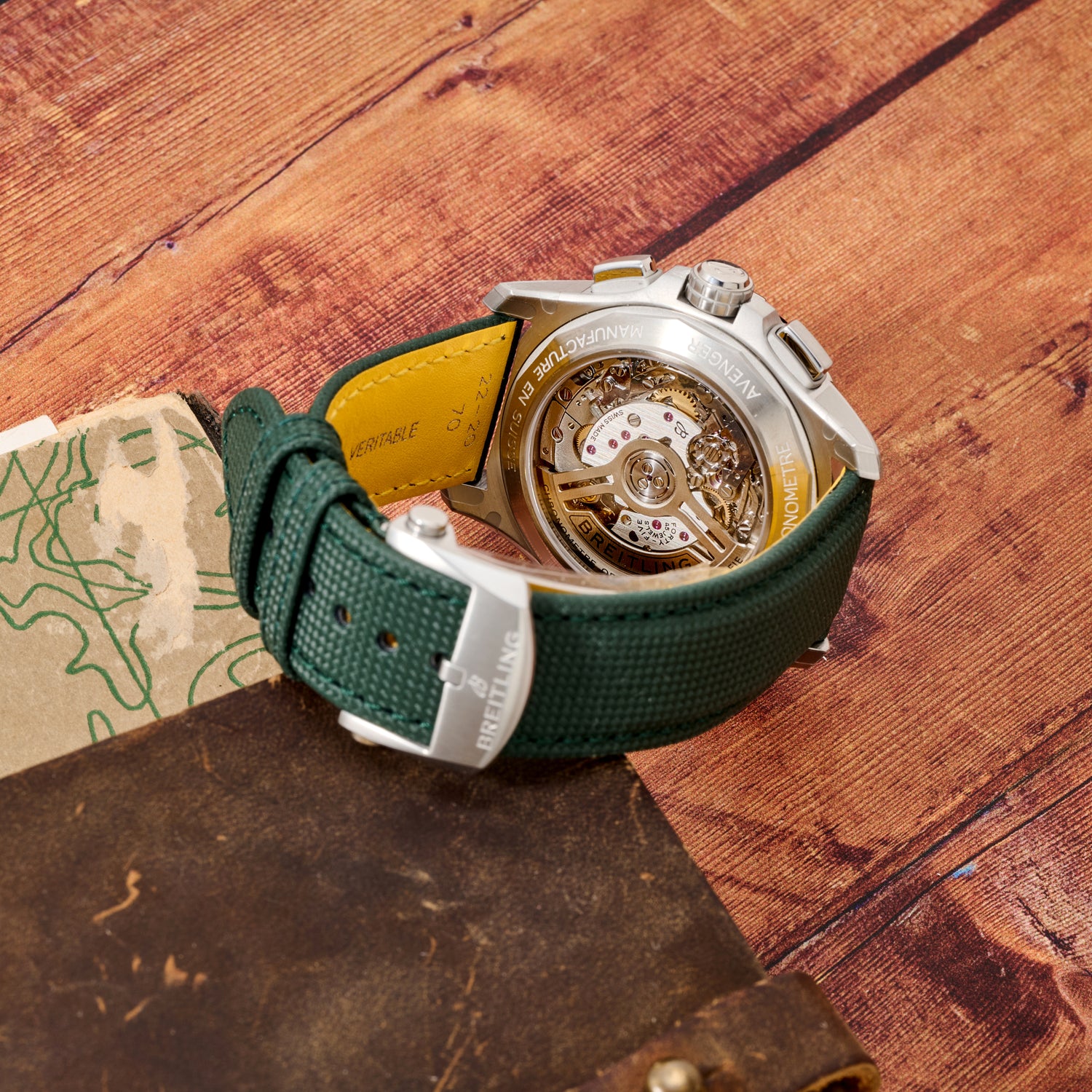 A Breitling Avenger B01 Chronograph 42 by Breitling, featuring a green strap, is shown face-down on a wooden surface to display its detailed exposed movement. A leather accessory and a partially visible map lie nearby.