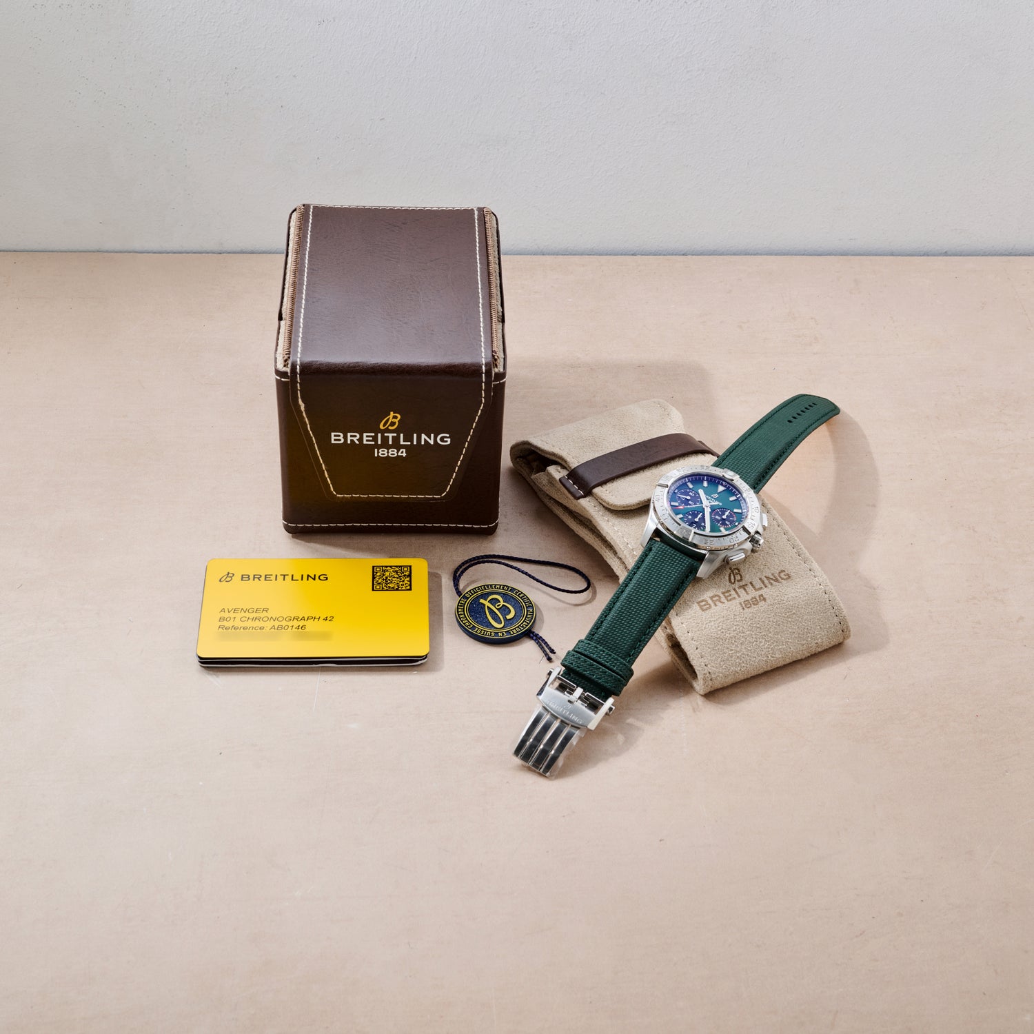 A Breitling Avenger B01 Chronograph 42 with a green strap, tan branded pouch, brown box, yellow warranty card, and round tag from Breitling are neatly arranged on a light beige surface.