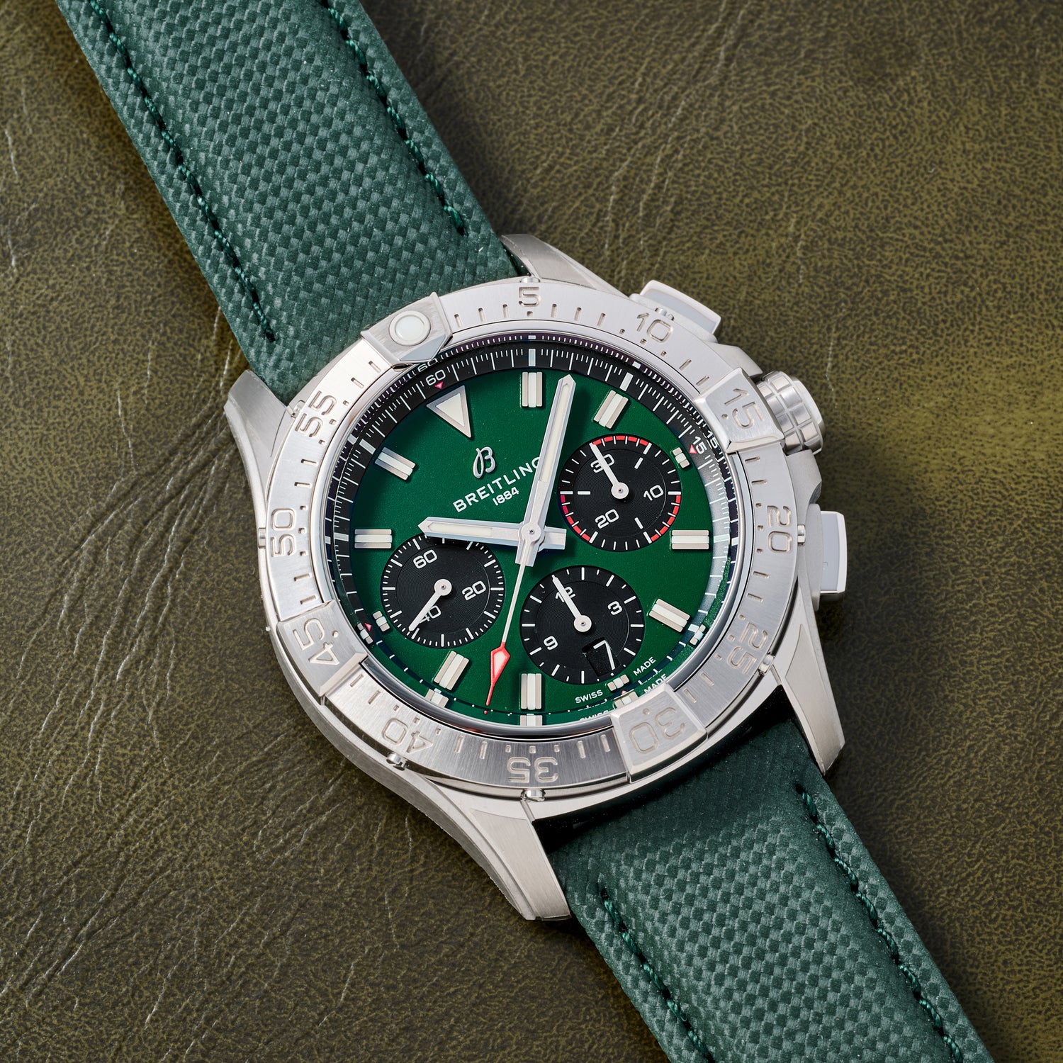A Breitling Avenger B01 Chronograph 42 with a green dial, three black subdials, silver bezel, and a matching green textured strap is displayed on an olive green leather surface.