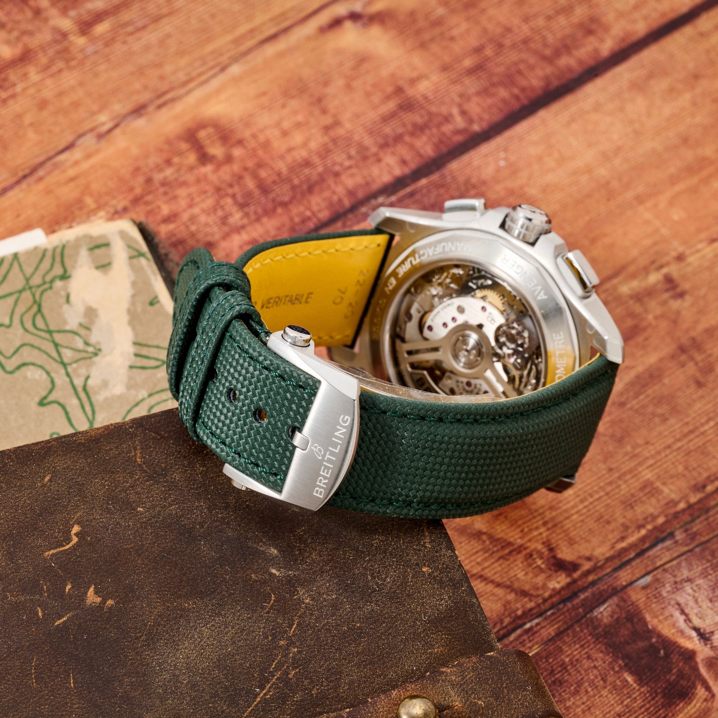 A close-up of the Breitling Avenger B01 Chronograph 42 watch with a green strap and open caseback showcasing its chronograph movement, displayed on weathered leather beside a map.