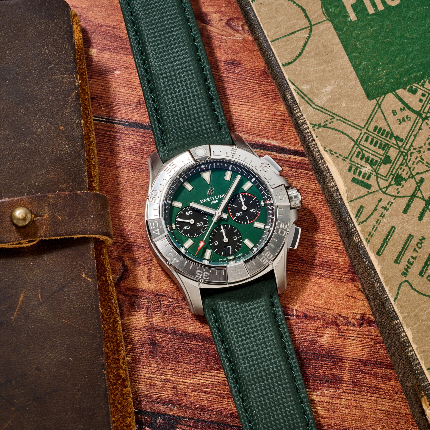 A Breitling Avenger B01 Chronograph 42 with a green strap and dial sits on a wooden surface next to a brown leather-bound notebook and a green map.