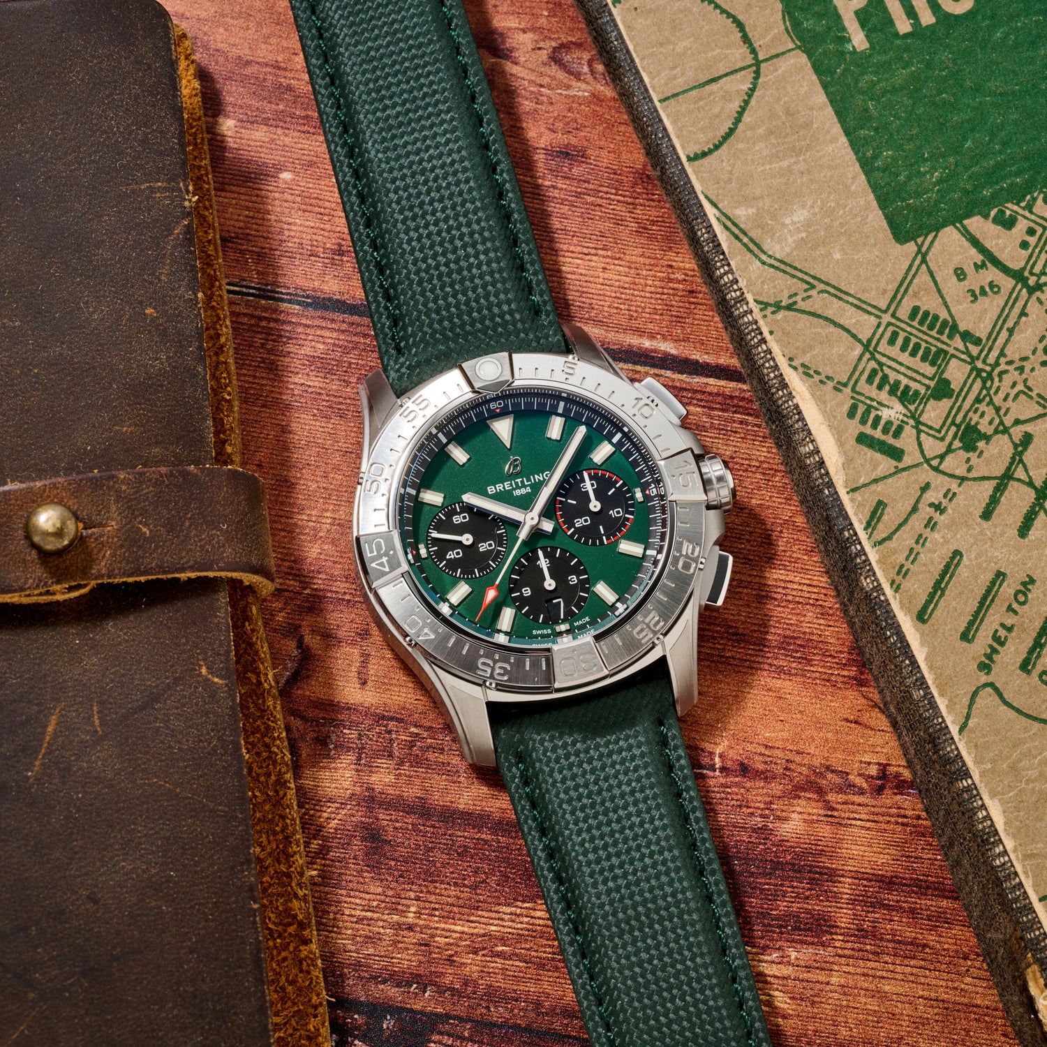 A Breitling Avenger B01 Chronograph 42 with a green strap and dial sits on a wooden surface next to a brown leather-bound notebook and a green map.