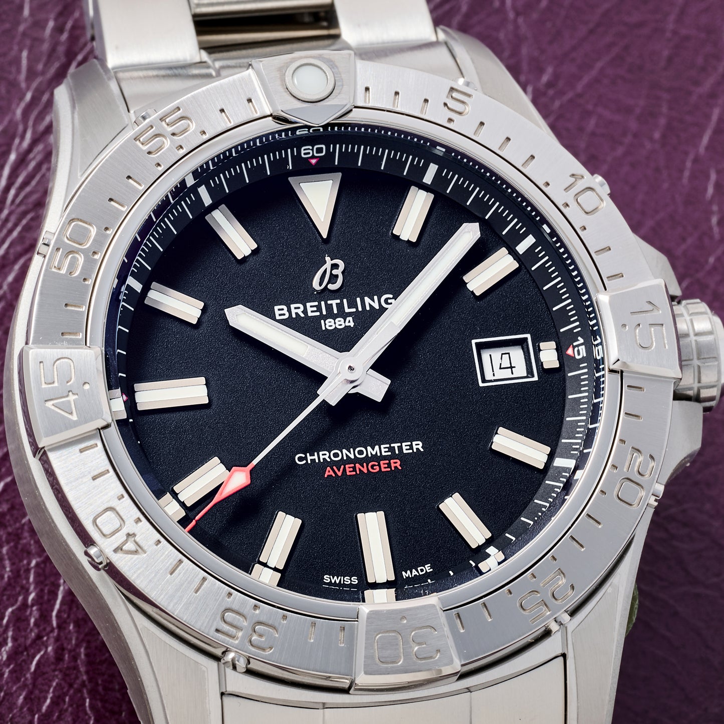 Close-up of the Breitling Avenger Automatic 42 by Breitling, featuring a stainless steel case and bracelet, black dial with white markers, date at 3 o’clock, and red-tipped second hand, set on a purple textured surface.