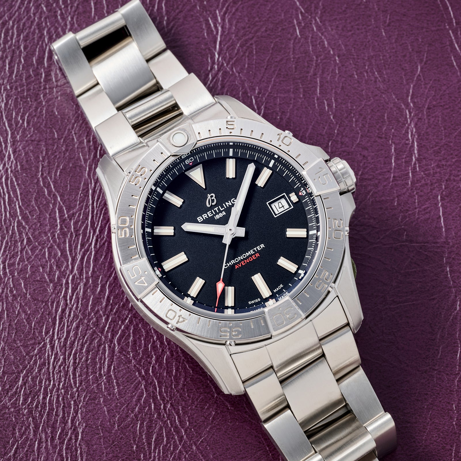 A Breitling Avenger Automatic 42 watch with a black dial, silver hour markers, and date window is shown on a purple textured leather surface. This stainless steel Breitling timepiece features a metal link bracelet.