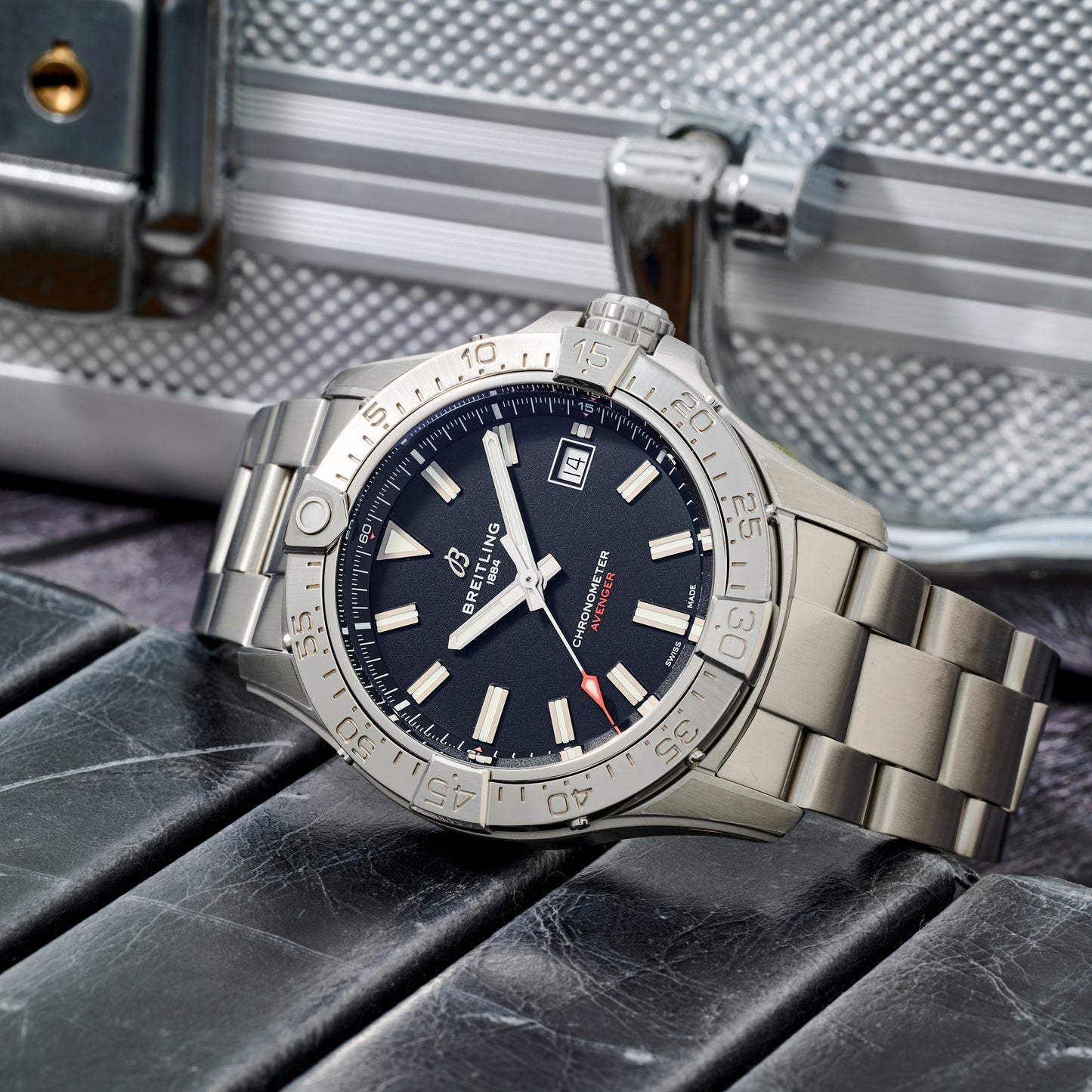 A Breitling Avenger Automatic 42 aviation watch with a stainless steel case, black dial, and silver bezel is displayed on a black leather surface. The watch shows 10:08 and the date as the 3rd, with a metallic case in the background.