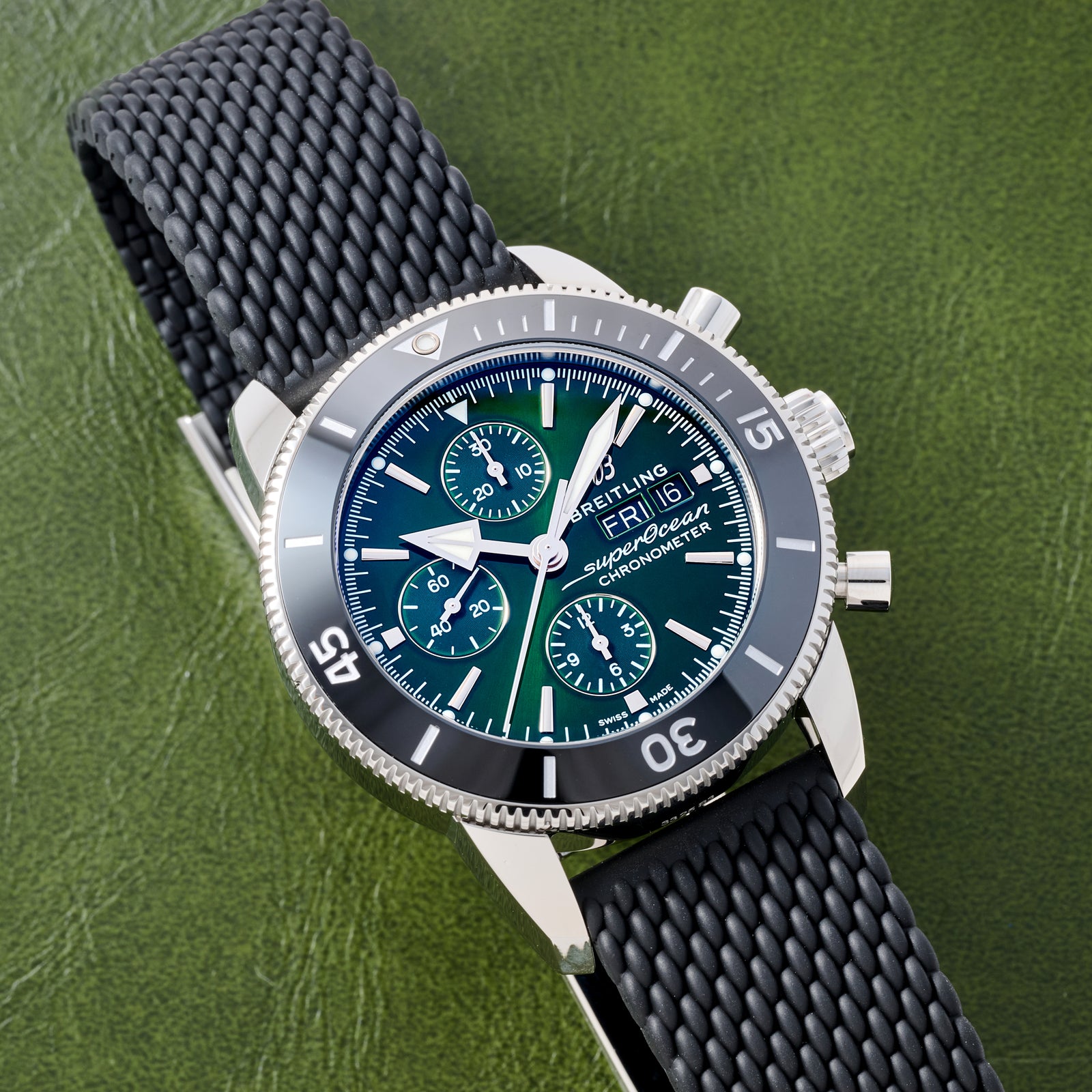 A close-up of the Breitling SuperOcean Heritage Chronograph 44 by Breitling, featuring a black textured strap, green and black bezel, three subdials, and white markers resting on a green textured surface.