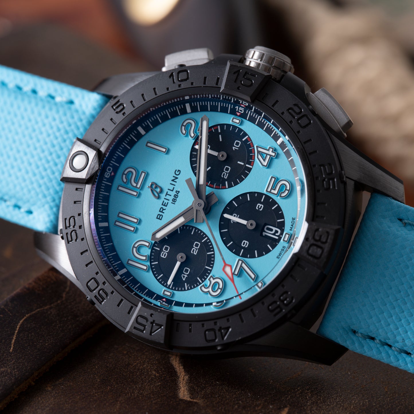 A close-up of a Breitling Super Avenger B01 Chronograph 42 wristwatch featuring a black bezel, light blue dial with three black subdials, bold Arabic numerals, and a textured light blue strap.