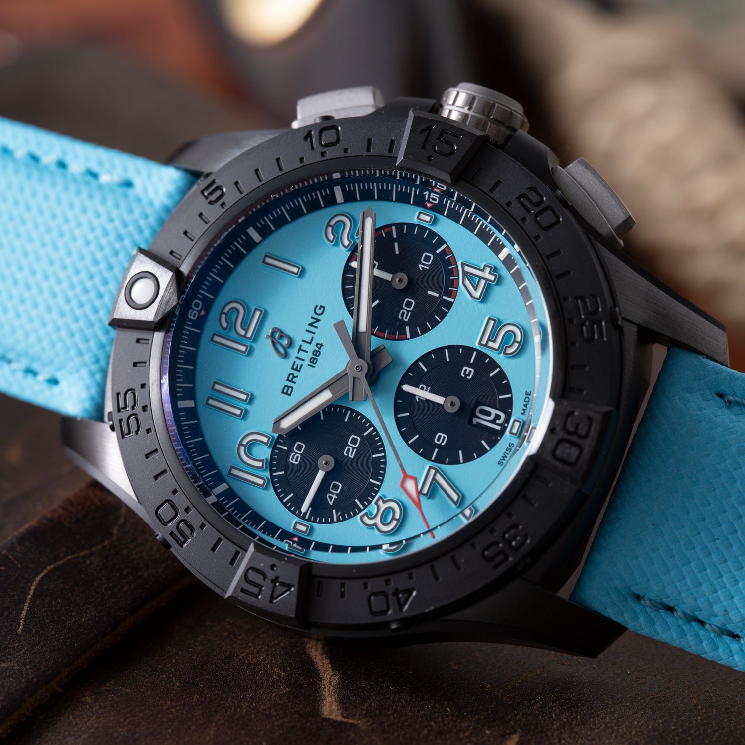 A close-up of a Breitling Super Avenger B01 Chronograph 42 wristwatch featuring a black bezel, light blue dial with three black subdials, bold Arabic numerals, and a textured light blue strap.