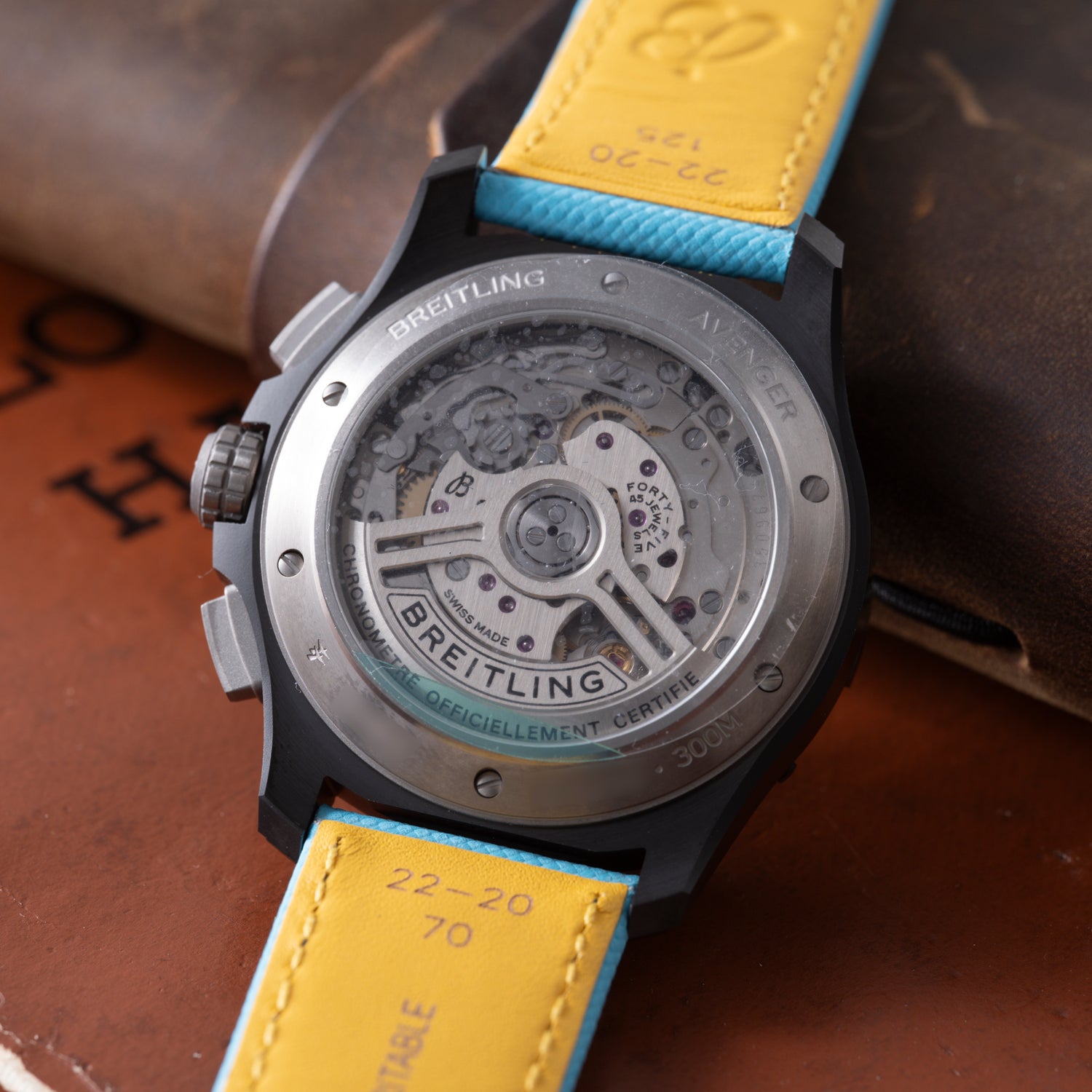 Close-up of the back of a Breitling Super Avenger B01 Chronograph 42, showing its detailed automatic movement through a transparent case back. The watch features a yellow and blue leather strap and is displayed on a brown surface.
