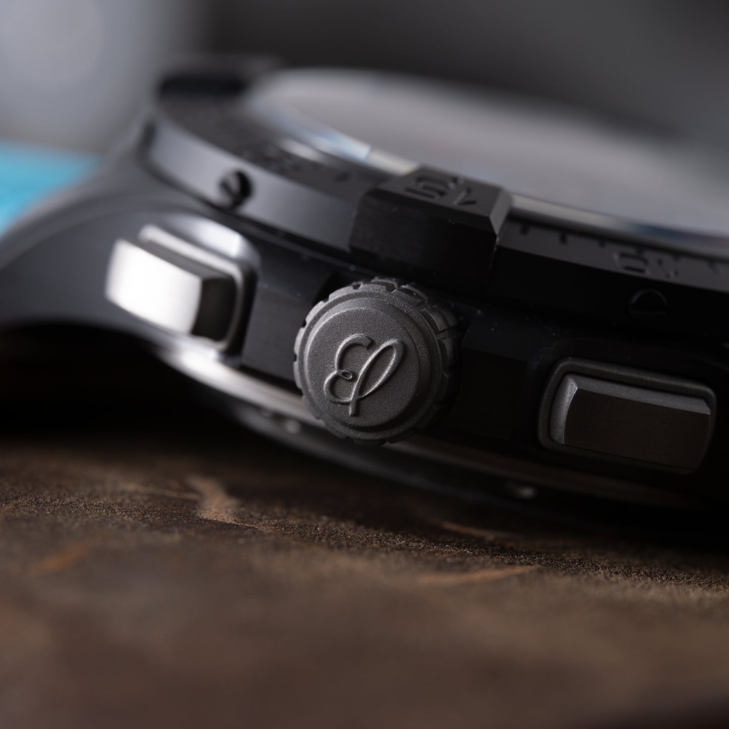 Close-up of a Breitling Super Avenger B01 Chronograph 42 crown with decorative engraving, flanked by two pushers, resting on brown textured material; details of the chronograph’s case and bezel are visible.