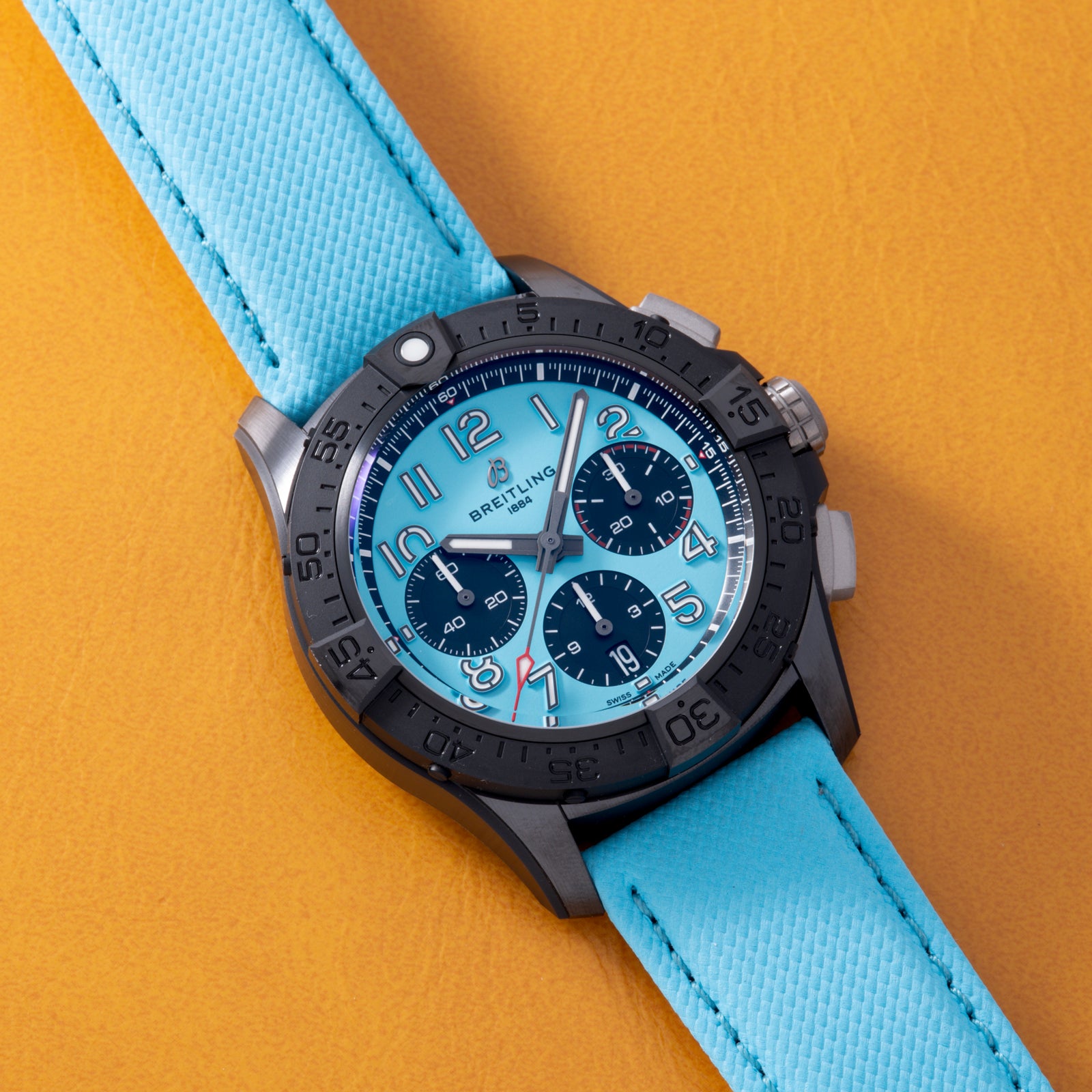 A Breitling Super Avenger B01 Chronograph 42 with a turquoise dial, black subdials, silver accents, and a bold black bezel on a matching strap is displayed on orange leather.