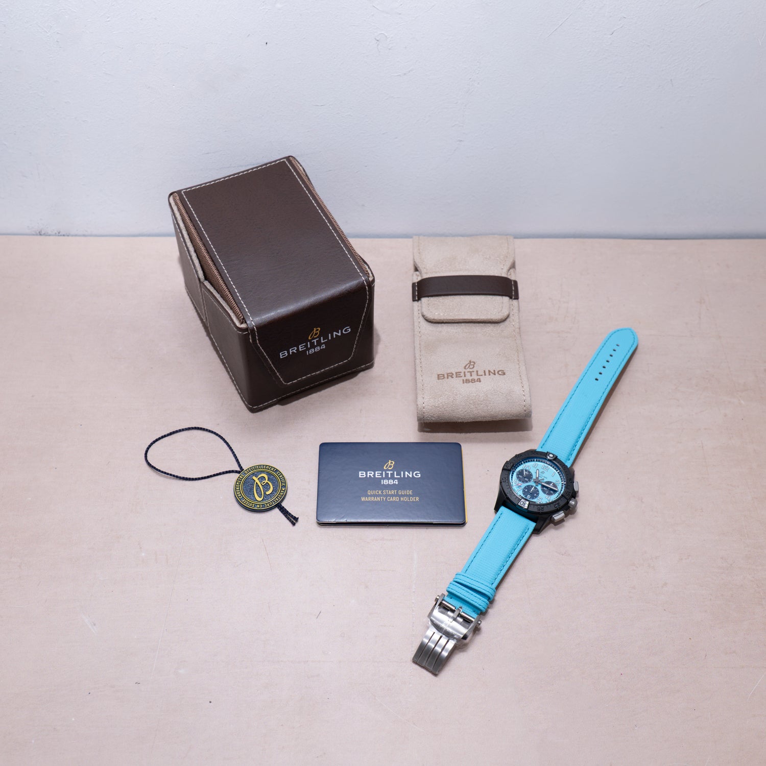 A Breitling Super Avenger B01 Chronograph 42 with a bright blue strap is displayed on a surface with a brown watch box, beige case, tag, and Breitling authentication card.