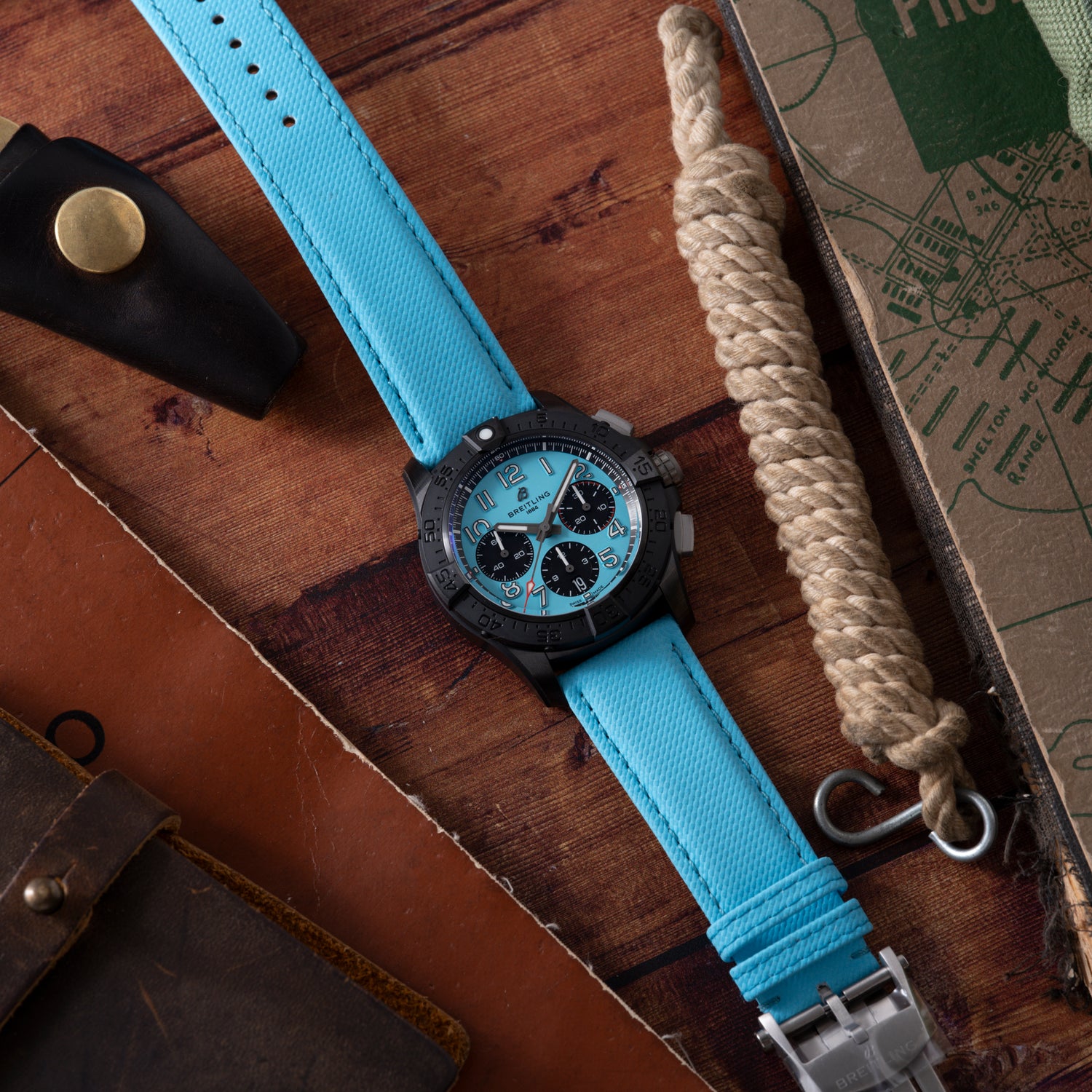 A Breitling Super Avenger B01 Chronograph 42 with a bright blue strap and black case rests on a wooden surface, alongside a sheathed knife, coiled rope, leather pouch, and part of a map.