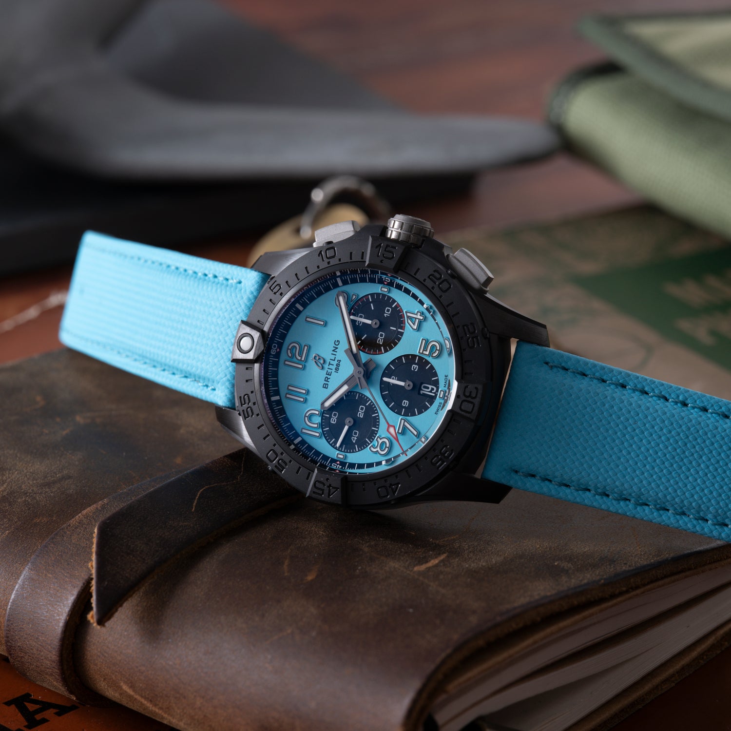 A black and blue Breitling Super Avenger B01 Chronograph 42 with a bright blue strap sits on a brown leather notebook, while a green pouch and papers are blurred in the background.