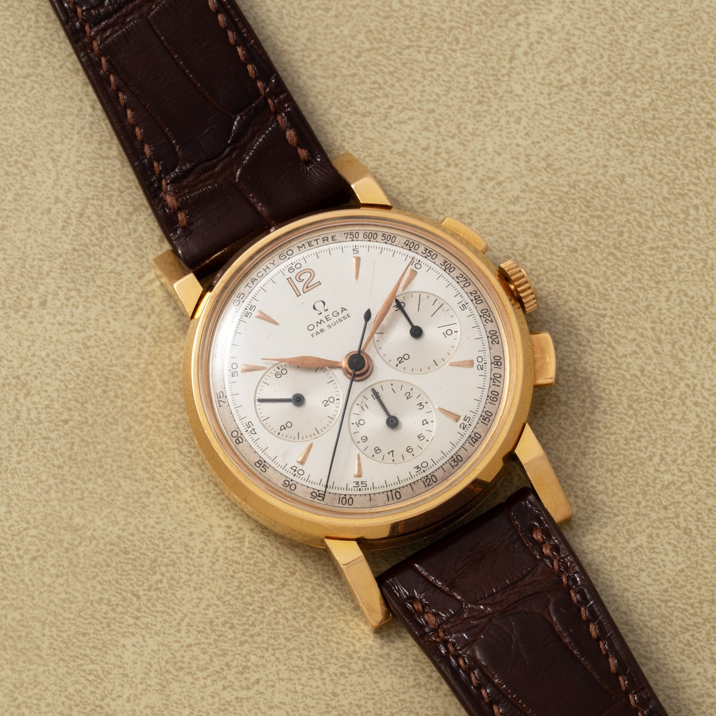 An Omega French Market Chronograph 'Calibre 321' wristwatch with a gold case, white dial, three subdials, and a brown leather strap is displayed on a textured beige surface.