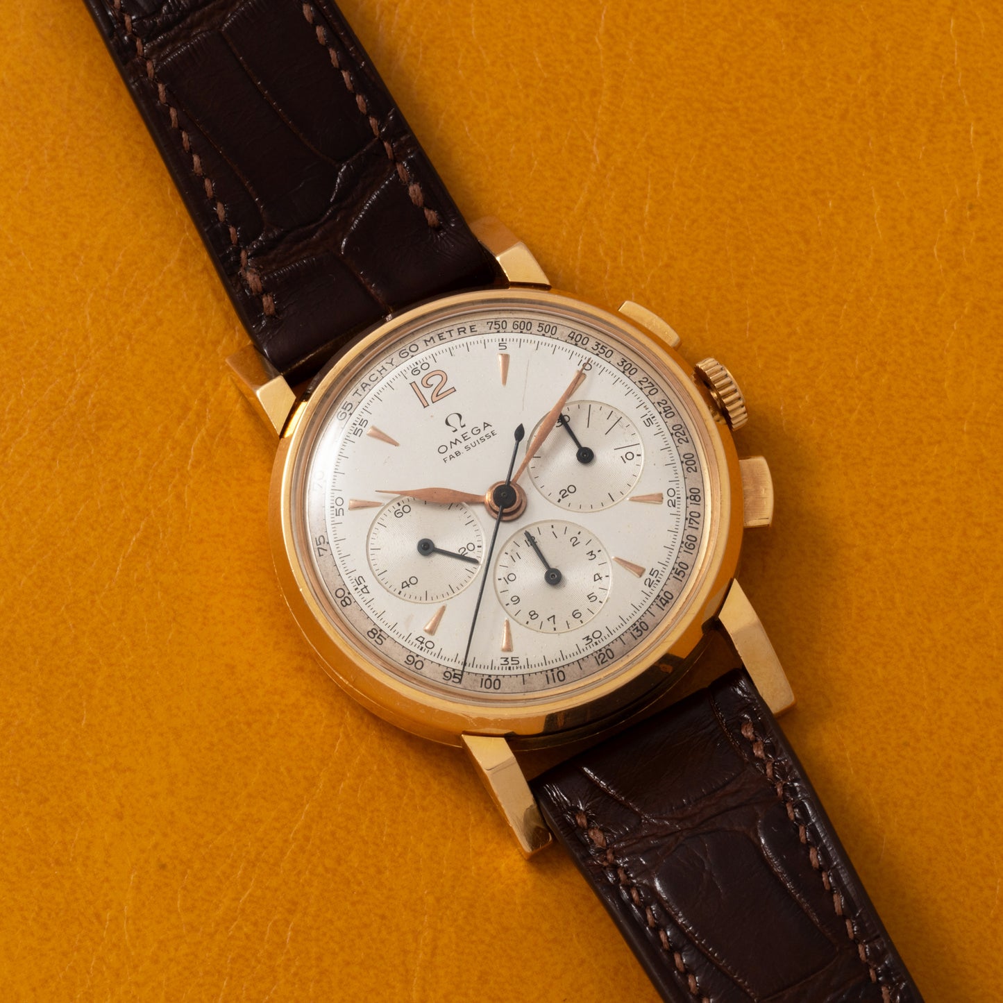 An Omega French Market Chronograph 'Calibre 321' with a gold case, white dial, brown leather strap, three subdials, and gold hour markers rests on a textured mustard yellow surface. It features the renowned Lemania Calibre 321 movement.