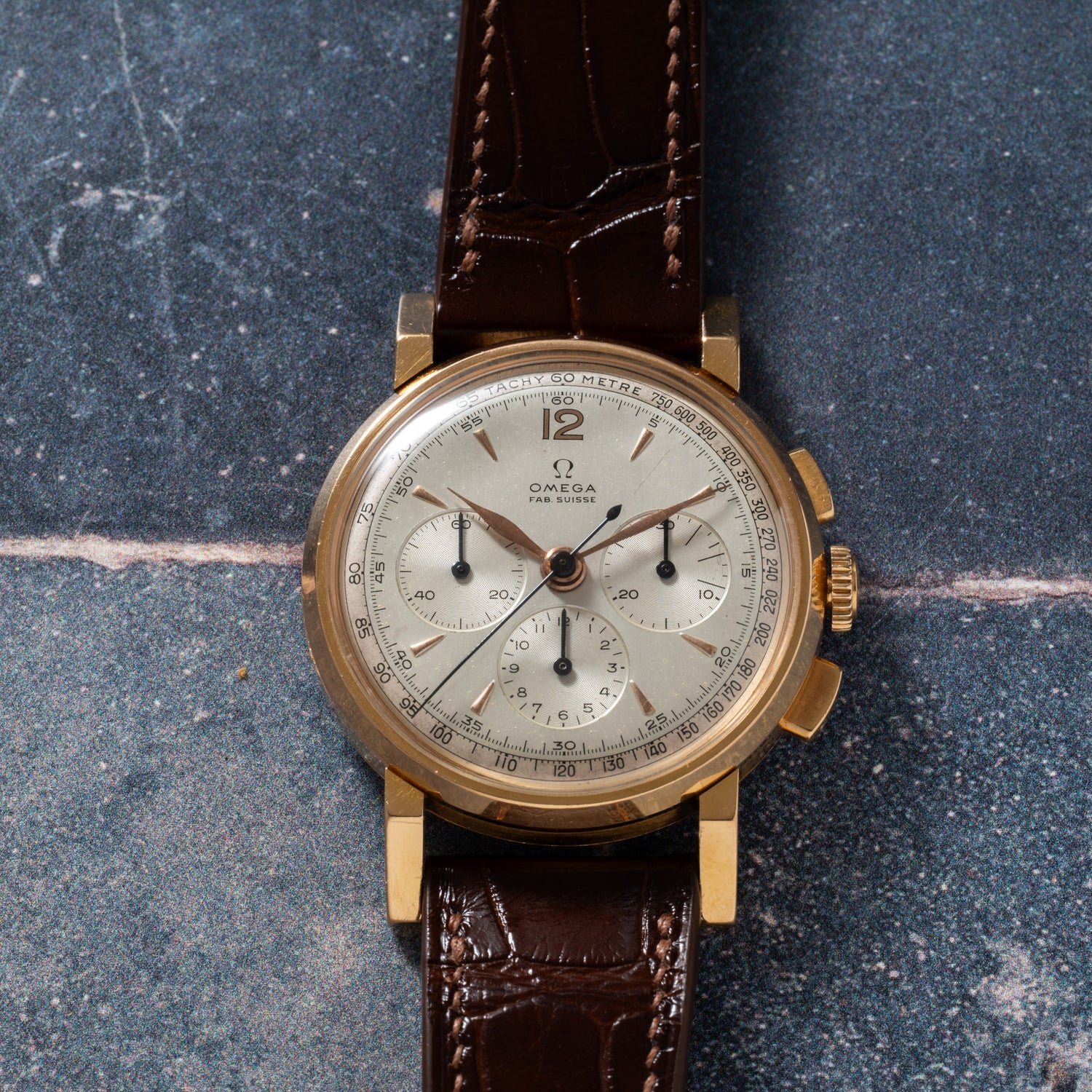 The Omega French Market Chronograph 'Calibre 321' features a gold case, brown leather strap, silver dial with three subdials and black numerals, powered by the Lemania Calibre 321, displayed on a textured blue-gray surface.