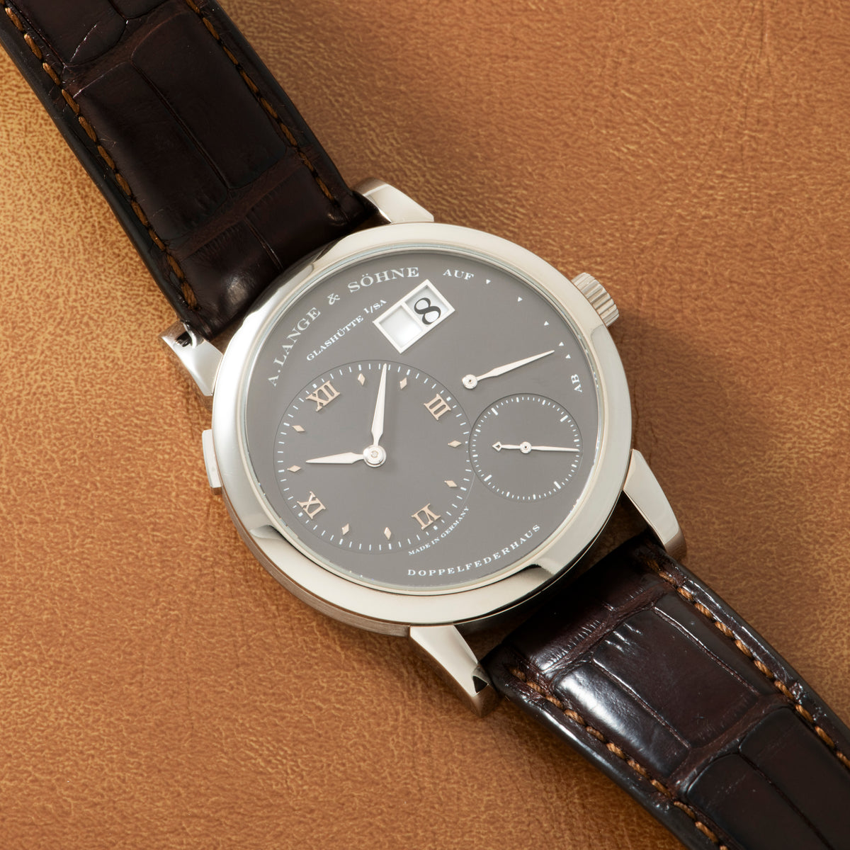 Silver-tone watch with gray dial, date window, Roman numerals, and dark brown leather strap on tan background.