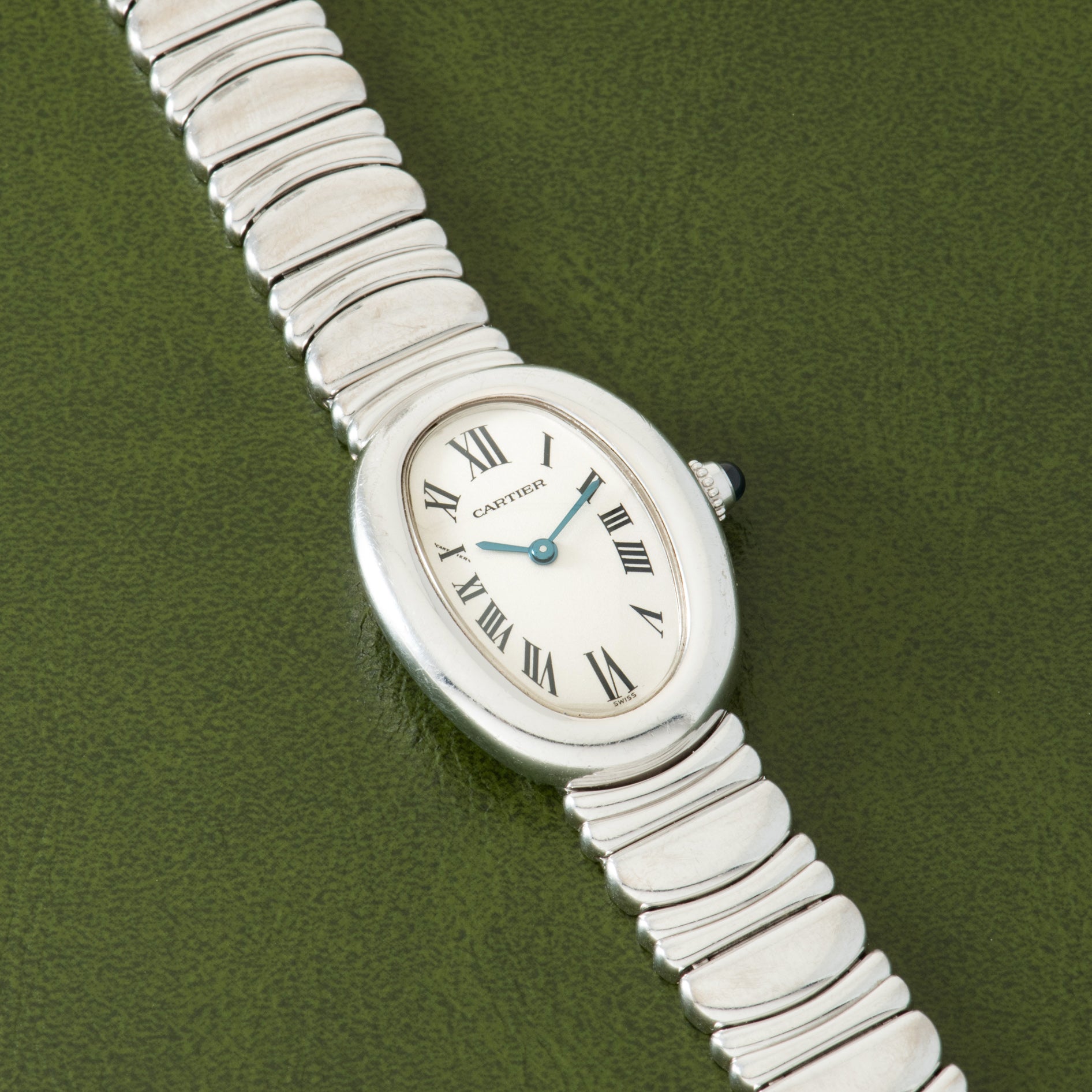 Silver-toned Cartier Baignoire watch with oval cream dial, blue hands, Roman numerals, and segmented metal bracelet on green 