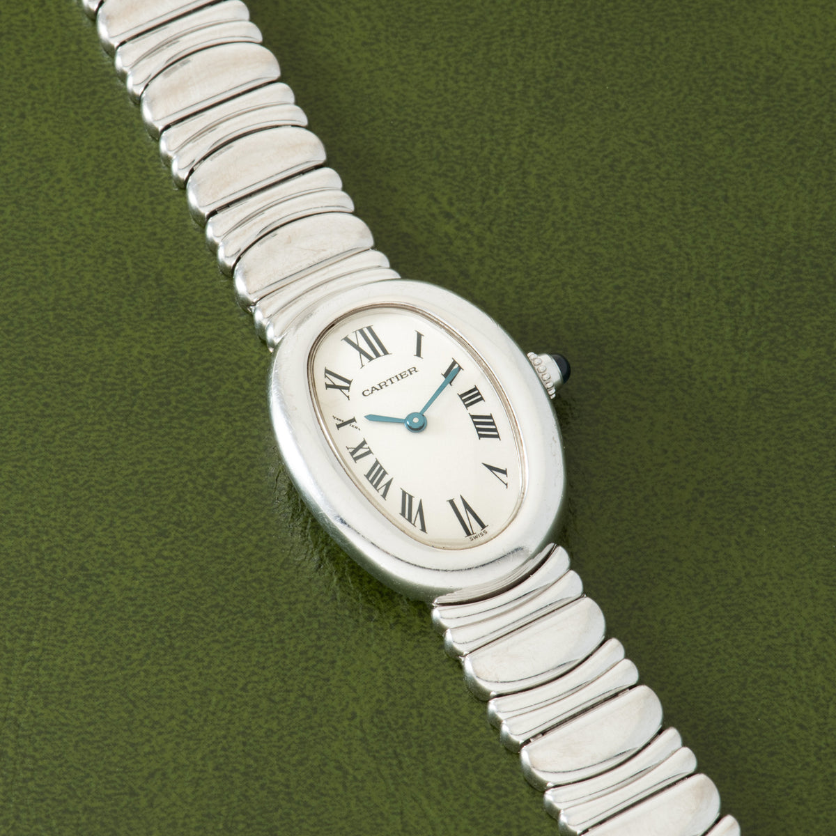 Silver-toned Cartier Baignoire watch with oval cream dial, blue hands, Roman numerals, and segmented metal bracelet on green 