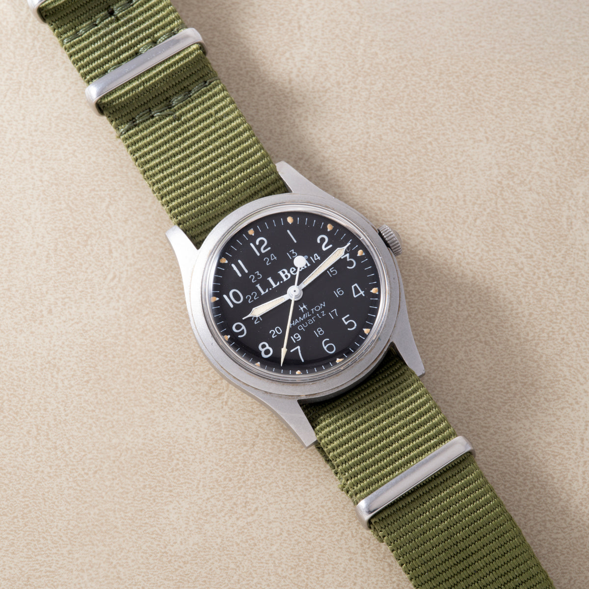 Stainless steel watch with black dial and olive green fabric strap with white hardware.