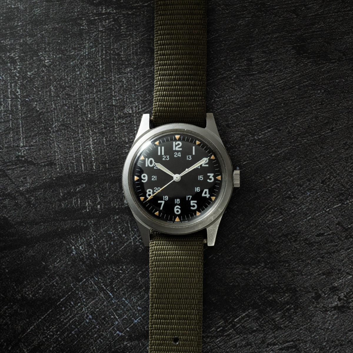 Stainless steel watch with black dial, 24-hour numerals, white hands, and olive green fabric strap on textured background.
