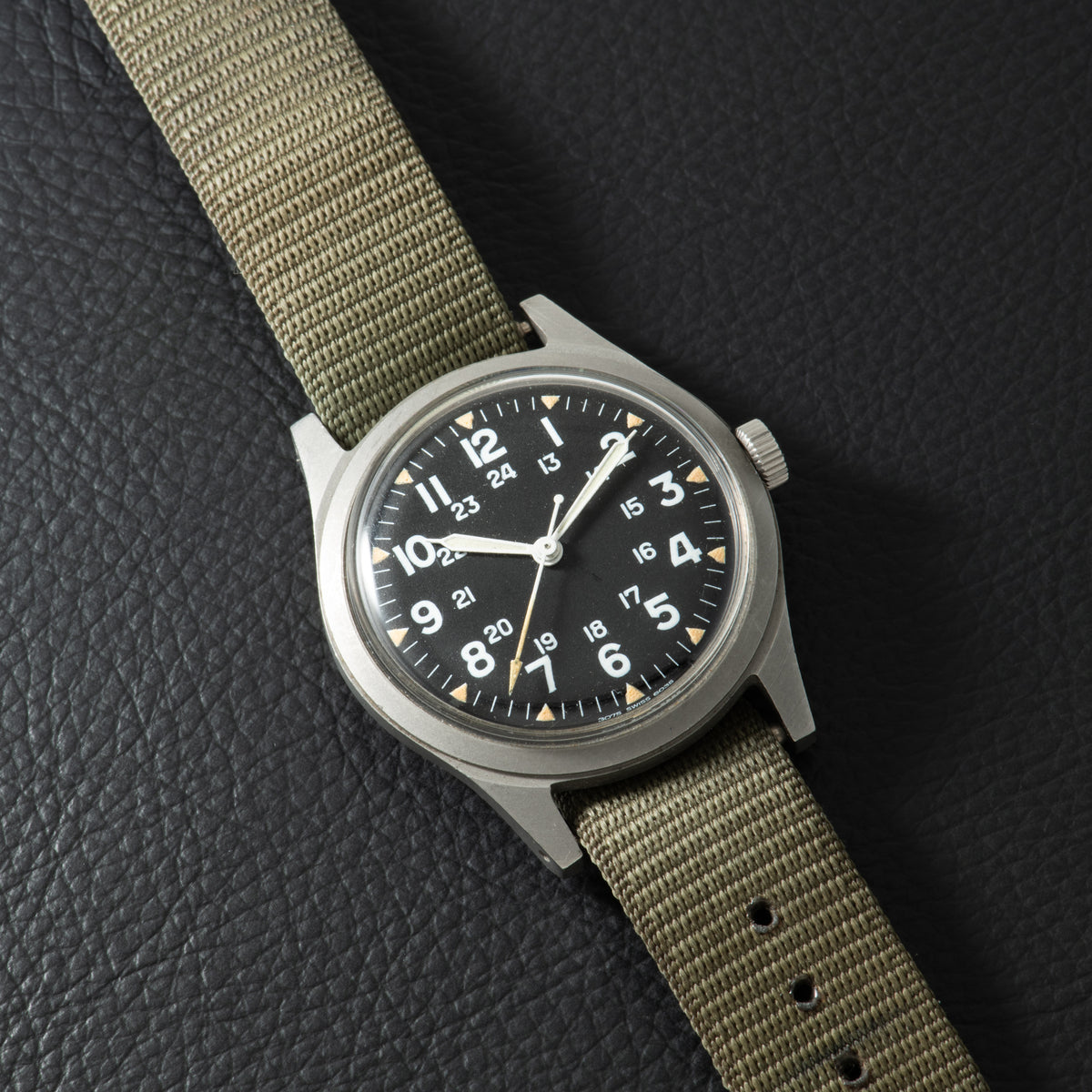 Stainless steel watch with black dial, Arabic numerals, white hands, and tan fabric strap.