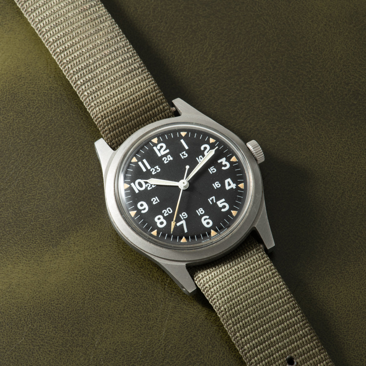 Hamilton GG-W-113 stainless steel watch with black dial, Arabic numerals, and tan fabric strap.