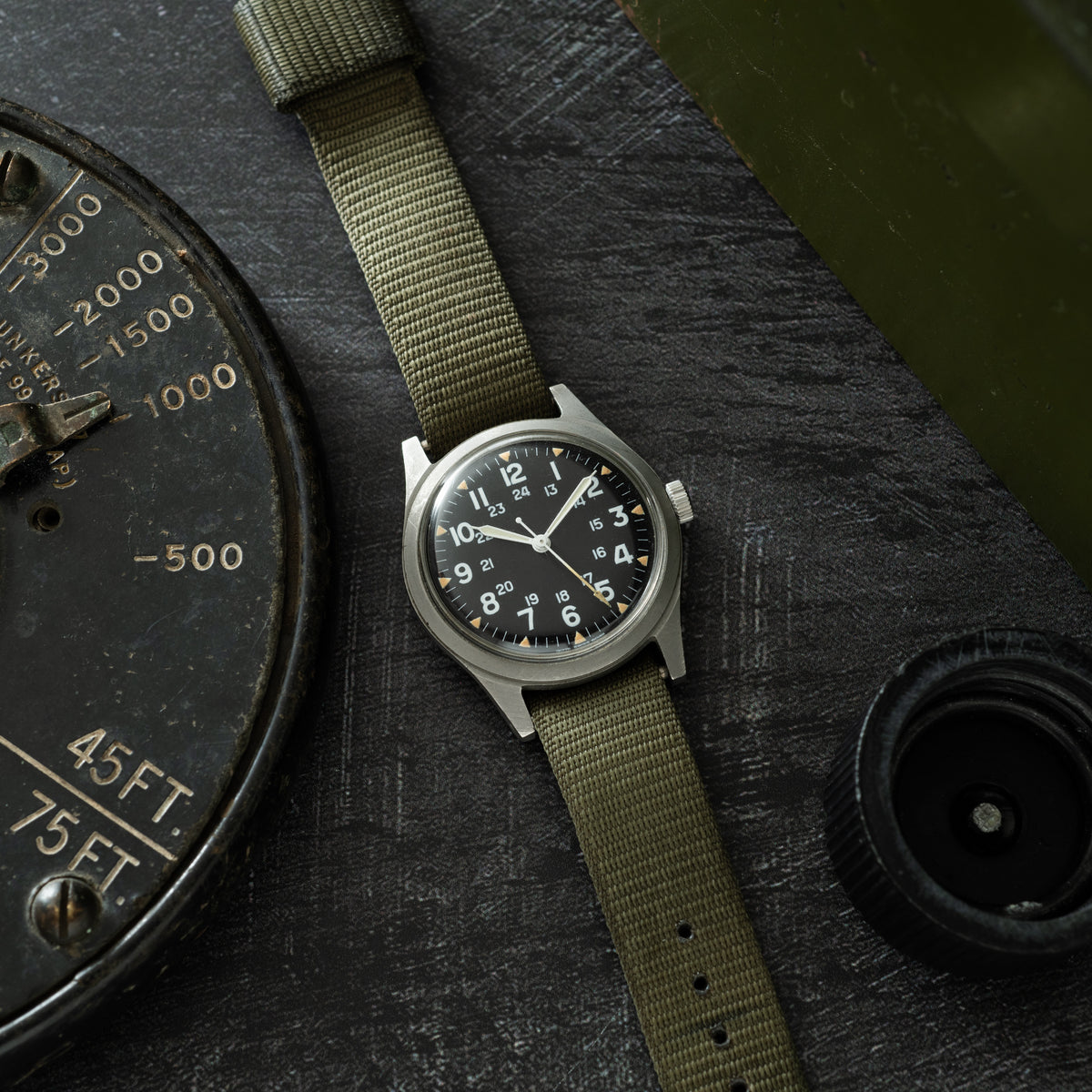 Stainless steel watch with black dial and white hands on olive green fabric strap, displayed with vintage gauge.