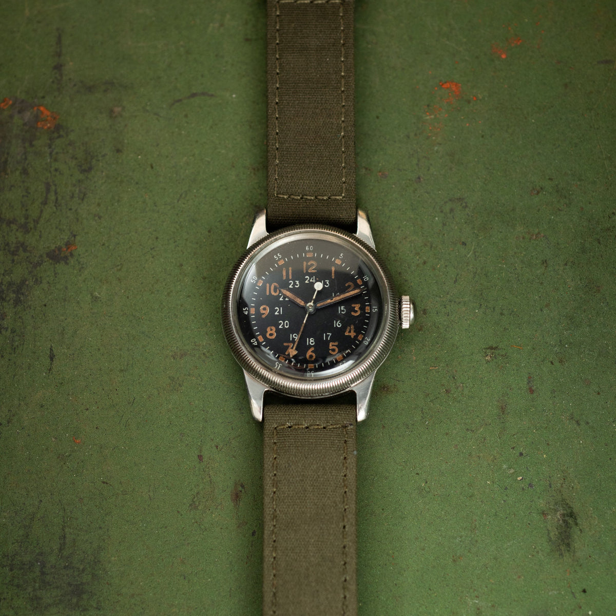 Waltham A-17 military watch with black dial, orange numerals, and olive canvas strap on green textured background.