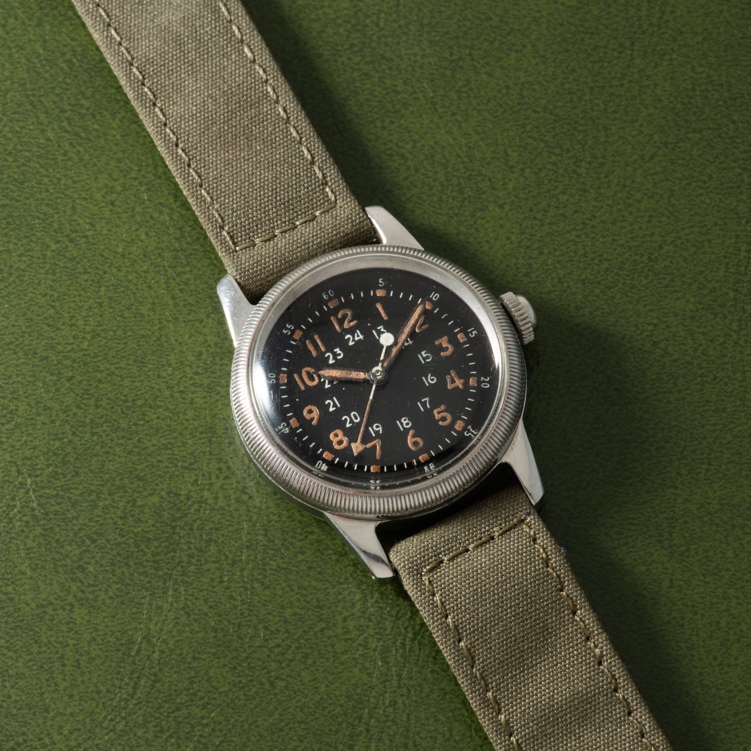 Waltham A-17 watch with black dial, Arabic numerals, and canvas strap on green background.