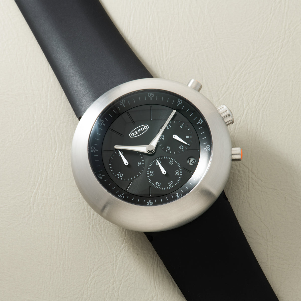 Ikepod Chronopod chronograph watch with silver case, black dial, and black leather strap on cream background.